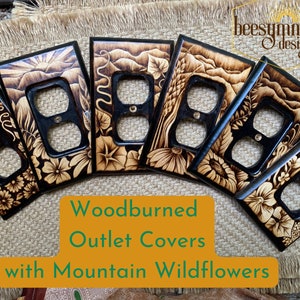 May include: A collection of woodburned outlet covers with mountain wildflower designs. Each cover features a black frame and a detailed, naturalistic wood-burned image of flowers, landscapes, and sunrises. The text "Woodburned Outlet Covers with Mountain Wildflowers" is visible.