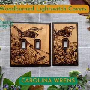 May include: Two woodburned light switch covers featuring a Carolina Wren perched on a branch with a nest of eggs. The covers are made of light and dark wood with a natural finish.