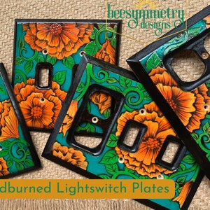 May include: Four woodburned light switch plates with a teal background and orange flowers. The plates are decorated with a floral design and have a black border.