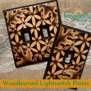 May include: Two woodburned light switch plates with a floral and leaf design. The plates are dark brown with a light brown woodgrain pattern. The text "beesymmetry designs" is visible in the top right corner of the image.