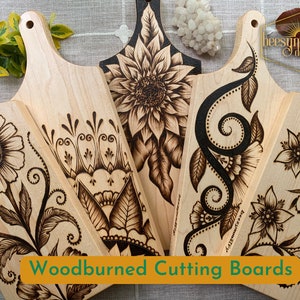 May include: Four wood-burned cutting boards with floral designs. The boards are made of light wood and have a natural finish. The designs are intricate and detailed, and they are all different. The boards are perfect for serving cheese, crackers, or other snacks.