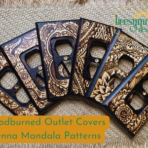 May include: Five woodburned outlet covers with intricate henna mandala patterns. The covers are arranged in a fan shape, with the top cover slightly overlapping the others. The wood is a dark brown color, and the patterns are a lighter brown color. The text "beesymmetry designs" is visible in the top right corner of the image.