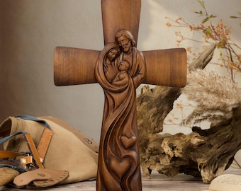 Peut inclure: Une croix en bois brun présentant un relief sculpté représentant une famille de trois personnages enlacés. Le motif comprend des lignes fluides qui se prolongent vers le bas en deux formes de cœur à la base de la croix.