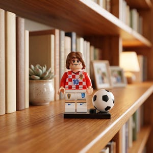 May include: A plastic minifigure of a soccer player with brown hair, wearing a red and white checkered jersey with the number 10 and matching white shorts. The figure stands on a black base next to a small black and white soccer ball.