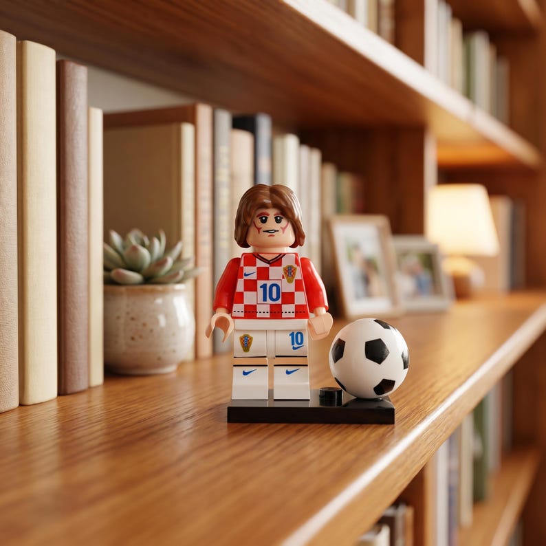 May include: A plastic minifigure of a soccer player with brown hair, wearing a red and white checkered jersey with the number 10 and matching white shorts. The figure stands on a black base next to a small black and white soccer ball.