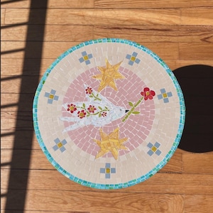 Può includere: Un piano d'appoggio circolare in mosaico raffigurante un uccello bianco centrale con motivi floreali rossi sulle ali, che tiene un fiore rosso nel becco. L'uccello è circondato da due stelle gialle su sfondo rosa, con un bordo di tessere blu.