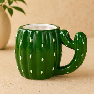 Op de afbeelding: Een glanzende donkergroene keramische mok in de vorm van een cactus met verticale ribbels en kleine wit geschilderde stippen die stekels voorstellen. Het handvat is gevormd als een gebogen cactusarm aan de zijkant van de beker.