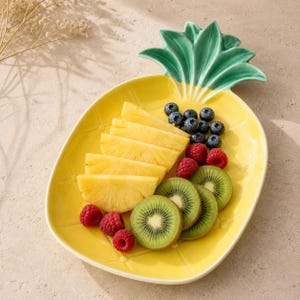 Puede incluir: Un plato de servicio de cerámica amarillo con forma de piña y una parte superior de hojas verdes, lleno de rodajas de piña, frambuesas frescas, arándanos y rodajas de kiwi. El plato presenta una superficie texturizada que imita la piel de una piña.