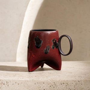 May include: A deep red ceramic mug with a speckled finish and irregular black glazed patches on the exterior. The mug features a rounded black handle and stands on three short, pointed feet, with a smooth black interior lining the rim.