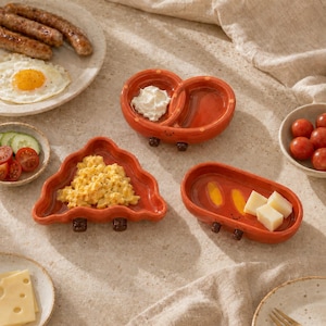 Puede incluir: Tres platos de cerámica naranja con forma de pretzel, triángulo y óvalo, cada uno con pies pequeños y una cara sonriente sencilla. Los platos contienen crema blanca, huevos revueltos y cubos de queso, dispuestos con alimentos de desayuno.