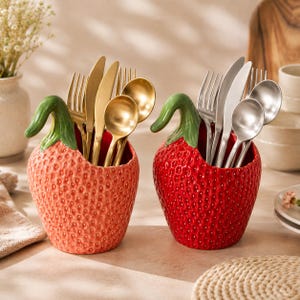 May include: Two ceramic strawberry-shaped utensil holders, one in a light coral and one in a deep red, each featuring a green stem handle. The coral holder contains gold-toned cutlery, while the red holder contains silver-toned cutlery.