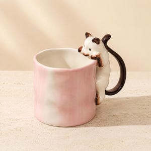 May include: A ceramic mug with a light pink and white marbled finish. The handle is shaped like a white cat with brown markings, appearing to climb over the rim of the mug. The cat has a long, curved dark brown tail that forms the handle grip.