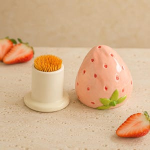 May include: A ceramic toothpick holder featuring a light pink strawberry-shaped lid with small red dots and green leaves. Beside the lid sits a plain white cylindrical base filled with a cluster of wooden toothpicks, accompanied by fresh strawberry slices.