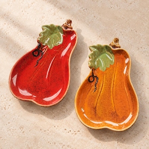 May include: Two ceramic dishes shaped like pumpkins, one in a vibrant red glaze and the other in a warm orange glaze. Each dish features a glossy finish, a sculpted green leaf detail at the top, and a small brown stem, with dark vine accents.