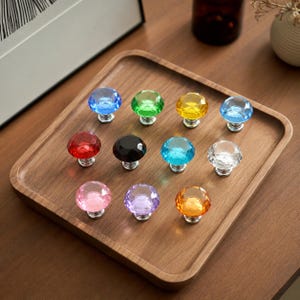 Könnte beinhalten: Eine Sammlung von elf facettierten, rautenförmigen Schrankknöpfen in verschiedenen durchscheinenden Farben wie Blau, Grün, Gelb, Rot, Schwarz, Türkis, Transparent, Rosa, Violett und Orange, jeweils mit einem silberfarbenen Metallsockel.