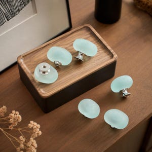 May include: Five oval-shaped cabinet knobs with a frosted, pale seafoam green finish and silver-toned metal bases. The knobs feature a smooth, rounded surface texture and are arranged in a cluster, showcasing their uniform design and colour.