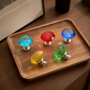 May include: Five faceted cabinet knobs with diamond-shaped heads in translucent colors including light blue, red, dark blue, yellow, and green. Each knob features a polished silver-tone metal base and a multi-faceted top surface that reflects light.