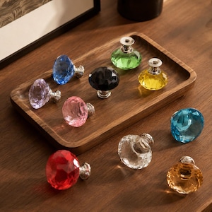 Pode incluir: Uma coleção de dez puxadores de armário facetados em forma de diamante em várias cores, incluindo vermelho, azul, amarelo, verde, rosa, roxo, preto, transparente e âmbar, cada um com uma base de metal prateado para fixação em móveis.