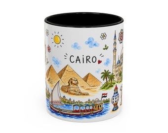 Pode incluir: Uma caneca de cerâmica branca com interior preto apresentando uma ilustração colorida em aquarela do Cairo. O design inclui as pirâmides, a Esfinge, um veleiro na água, uma mesquita e o texto CAIRO com um pequeno ícone de coração vermelho.