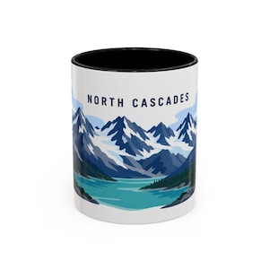 May include: A white ceramic mug with a black interior and rim, featuring a graphic illustration of the North Cascades mountains. The design displays snow-capped blue peaks, a turquoise lake, and dark green pine trees with the text NORTH CASCADES.