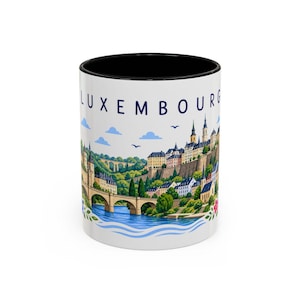 Può includere: Una tazza in ceramica bianca con interno e bordo neri, caratterizzata da un'illustrazione colorata del paesaggio urbano di Lussemburgo, inclusi ponti in pietra, edifici storici e un fiume, con il testo LUXEMBOURG stampato in blu scuro.