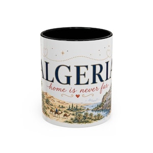Peut inclure: Un mug en céramique blanc avec un intérieur et un bord noirs. L'extérieur présente le texte ALGERIA en grandes lettres bleues, avec la phrase home is never far en écriture rouge. Un paysage désertique avec des chameaux est illustré.
