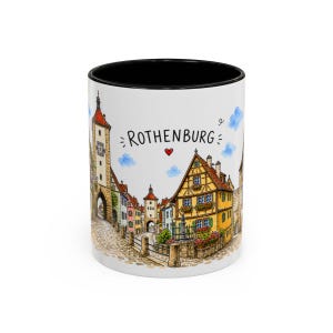 Peut inclure: Un mug en céramique blanche avec un intérieur noir, illustré d'une aquarelle colorée représentant l'architecture de la ville de Rothenburg, avec des maisons à colombages, une tour en pierre, des rues pavées, le texte ROTHENBURG et un cœur rouge.