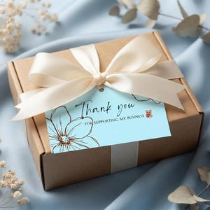 May include: A square brown cardboard gift box tied with a cream-coloured satin ribbon. A light blue rectangular tag is attached to the ribbon, featuring a black floral line drawing and the text Thank you FOR SUPPORTING MY BUSINESS.