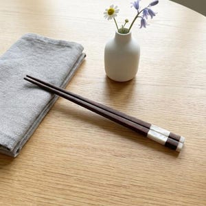 Op de afbeelding: Een paar donkerbruine houten eetstokjes met witte rechthoekige inlays aan de uiteinden, geplaatst naast een gevouwen grijze linnen servet en een kleine witte keramische vaas met een madeliefje en een blauw klokje.