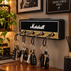 May include: A black Marshall amplifier-style wall-mounted key holder featuring a gold-coloured control panel with four input jacks. Four guitar plug keychains are inserted into the jacks, each holding a set of metal keys and car key fobs.