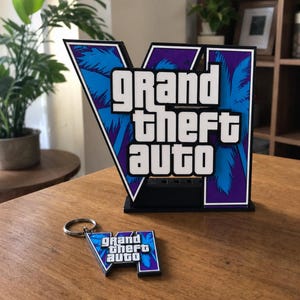 Op de afbeelding: Een 3D-geprint displaybord met het Grand Theft Auto VI-logo met witte letters op een paarse en blauwe achtergrond met palmbomen, vergezeld van een bijpassende miniatuur sleutelhangerversie van hetzelfde logo aan een metalen ring.
