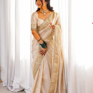 May include: A cream-colored saree featuring a wide, ornate gold border with intricate embroidery and sequin detailing. The garment includes a matching short-sleeved blouse with gold trim, paired with a gold choker necklace and dark green bangles.
