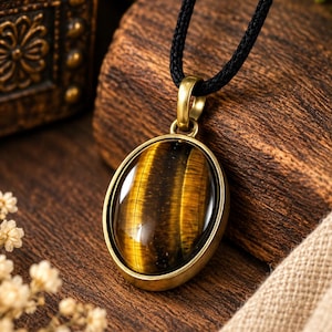 Pode incluir: Um pingente oval de pedra olho de tigre engastado numa moldura de metal cor de latão. A pedra lisa e polida apresenta bandas características de cor castanho-dourado e preto com efeito chatoyant. O pingente está suspenso num cordão preto.