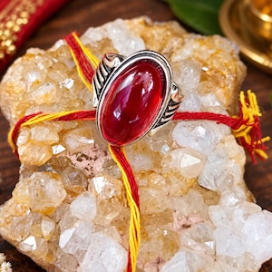 Puede incluir: Un anillo de plata con una piedra grande, lisa y de forma ovalada en color rojo intenso, montada en un marco metálico. El anillo está colocado sobre un grupo de cristales translúcidos de color amarillo pálido y blanco, atados con hilos finos entrelazados rojos y amarillos.