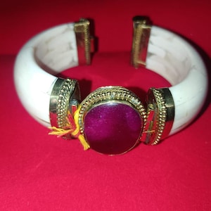 May include: A hinged cuff bracelet featuring a smooth white band accented with gold-toned metal hardware. A large, oval-shaped, deep magenta stone is set in the center within a decorative gold-toned frame, with a small yellow thread attached.