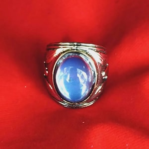 Può includere: Un anello in metallo color argento con una grande pietra cabochon ovale e liscia di colore blu incastonata al centro. La fascia dell'anello presenta dettagli intricati e testurizzati sui lati, che circondano l'incastonatura della pietra blu.