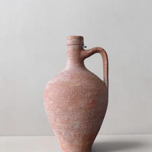 Könnte beinhalten: Ein Terrakotta-Krug mit einer verwitterten, strukturierten Oberfläche und mattem Finish. Das Gefäß hat einen bauchigen Körper, der sich zu einem schmalen Hals verjüngt, und ist mit einem einfachen Rand sowie einem geschwungenen Henkel versehen.