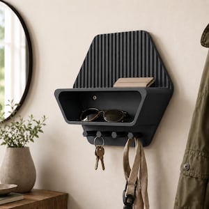 Puede incluir: Un organizador de pared negro mate con un panel trasero acanalado, un estante de almacenamiento horizontal y cuatro ganchos hexagonales. El estante sostiene un par de gafas de sol y una cartera pequeña, mientras que los ganchos muestran llaves y un cordón.