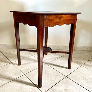 Peut inclure: Une table d'appoint carrée en bois avec une finition brun foncé, dotée de quatre pieds droits et d'un tablier festonné. La table comprend une petite étagère centrale circulaire positionnée entre les pieds, offrant une structure de base stable.