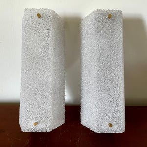 Könnte beinhalten: Ein Paar rechteckige Wandleuchten mit strukturierten, klaren Glasscheiben und einer rauen, kristallinen Oberfläche. Jede Leuchte ist mit zwei kleinen, runden, messingfarbenen Metallbefestigungen an den oberen und unteren Kanten befestigt.