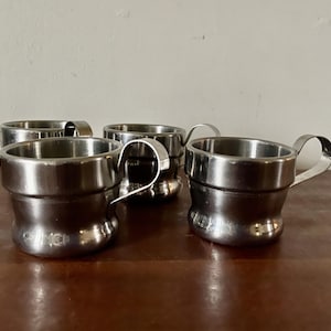 May include: A set of three small, polished stainless steel cups, each featuring a cylindrical body with a slightly flared rim and a curved metal handle attached to the side. The cups have a smooth, reflective metallic finish and a simple design.