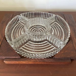Puede incluir: Un plato de servicio dividido de vidrio transparente con un compartimento circular central y cuatro secciones curvas circundantes, todas con un patrón de costillas verticales, colocado dentro de un marco de madera marrón oscuro con acentos geométricos.