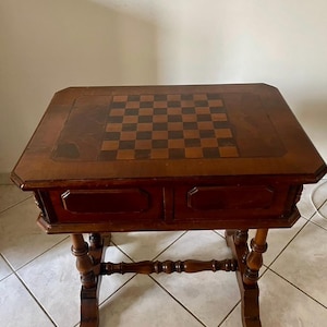 Peut inclure: Une table de jeu rectangulaire en bois dotée d'un échiquier intégré sur le dessus. La table est fabriquée en bois brun foncé et comprend deux tiroirs frontaux avec des panneaux rectangulaires, reposant sur quatre pieds tournés.