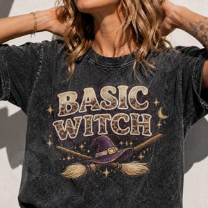 Può includere: Una t-shirt grigio antracite con effetto slavato che presenta la scritta BASIC WITCH in stampa leopardata. Sotto il testo, un cappello da strega viola poggia su due scope di legno incrociate, circondato da piccole grafiche di stelle e lune.