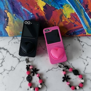 May include: Two foldable phone cases, one black and one pink, each featuring a raised heart-shaped grip on the back. Both cases include a matching beaded wrist strap with black and pink beads, small black bows, and circular charms attached to the base.