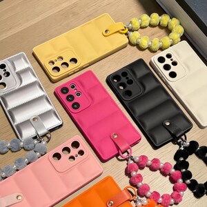 Puede incluir: Una colección de siete fundas rectangulares para smartphone con textura acolchada. Cada funda cuenta con un recorte para la cámara y una correa de muñeca a juego hecha de pompones difusos y cuentas blancas. Colores: amarillo, plata, rosa, negro, crema y naranja.
