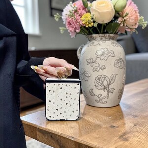 Può includere: Una custodia per telefono quadrata e pieghevole con sfondo color crema e un motivo ripetuto di piccoli fiocchi neri e cuori dorati. La custodia presenta una cornice nera e un anello circolare dorato fissato sul bordo superiore.