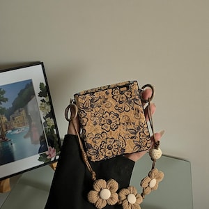 May include: A square cork crossbody bag featuring a black floral print. The bag includes a brown cord strap adorned with three-dimensional beige fabric flowers and a small round bead. The bag has a rectangular shape with rounded corners.