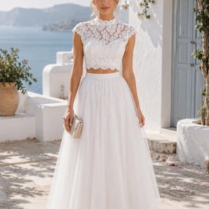 Puede incluir: Un conjunto blanco de dos piezas que consta de un top corto de encaje con cuello alto, mangas cortas y dobladillo festoneado, junto con una falda de tul fluida hasta el suelo. El top presenta detalles de encaje floral y la falda es de capas.