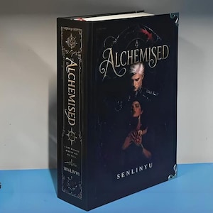 Op de afbeelding: Een hardcover boek getiteld Alchemised door Senlinyu met een zwarte kaft en een illustratie van twee figuren. De voorkant en rug tonen gouden letters en gedetailleerde decoratieve randen. Het boek is voorzien van zilverkleurige metalen hoekstukken.